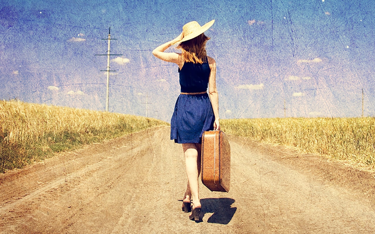 Eine attraktive Frau mit Reisekoffer auf einer Landstrasse im Vintage Stiel, Sie hat einen langen Rücken als Symbol für ein Leben ohne Rückenschmerzen